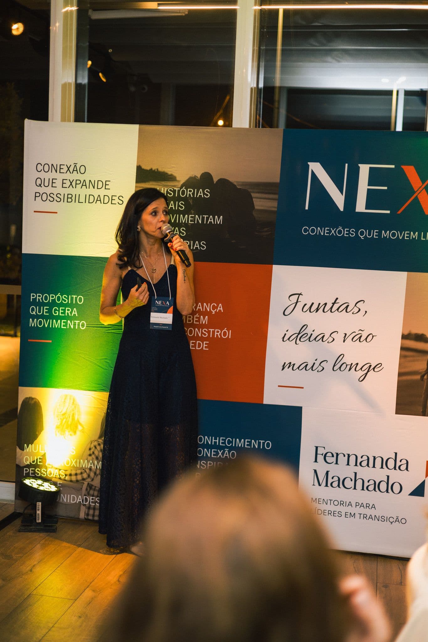 Fernanda Machado ao microfone, de pé ao lado do painel com o próprio nome, iluminada por luz quente.