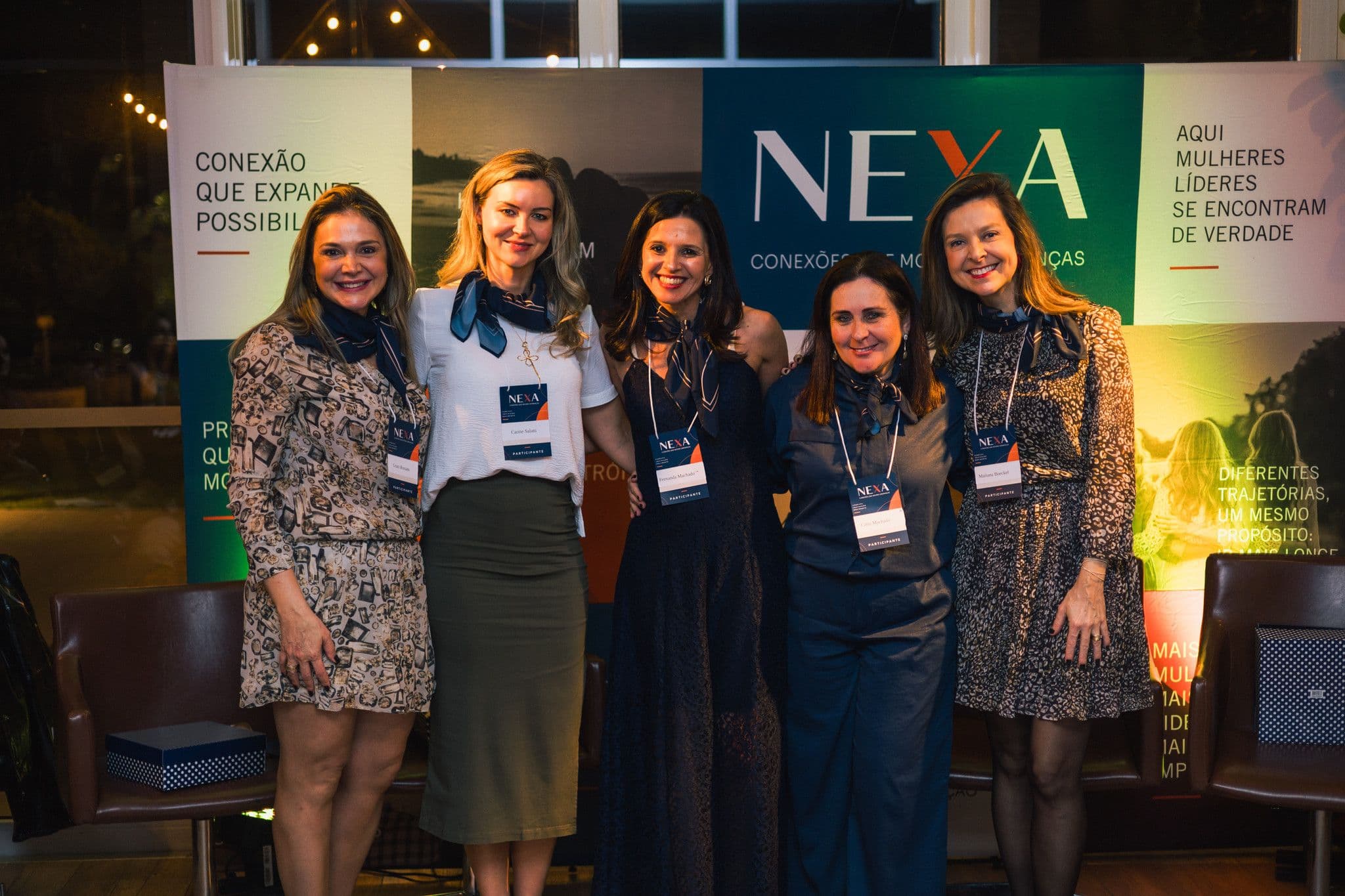 Cinco mulheres posam lado a lado diante do painel NEXA, com crachás no pescoço.