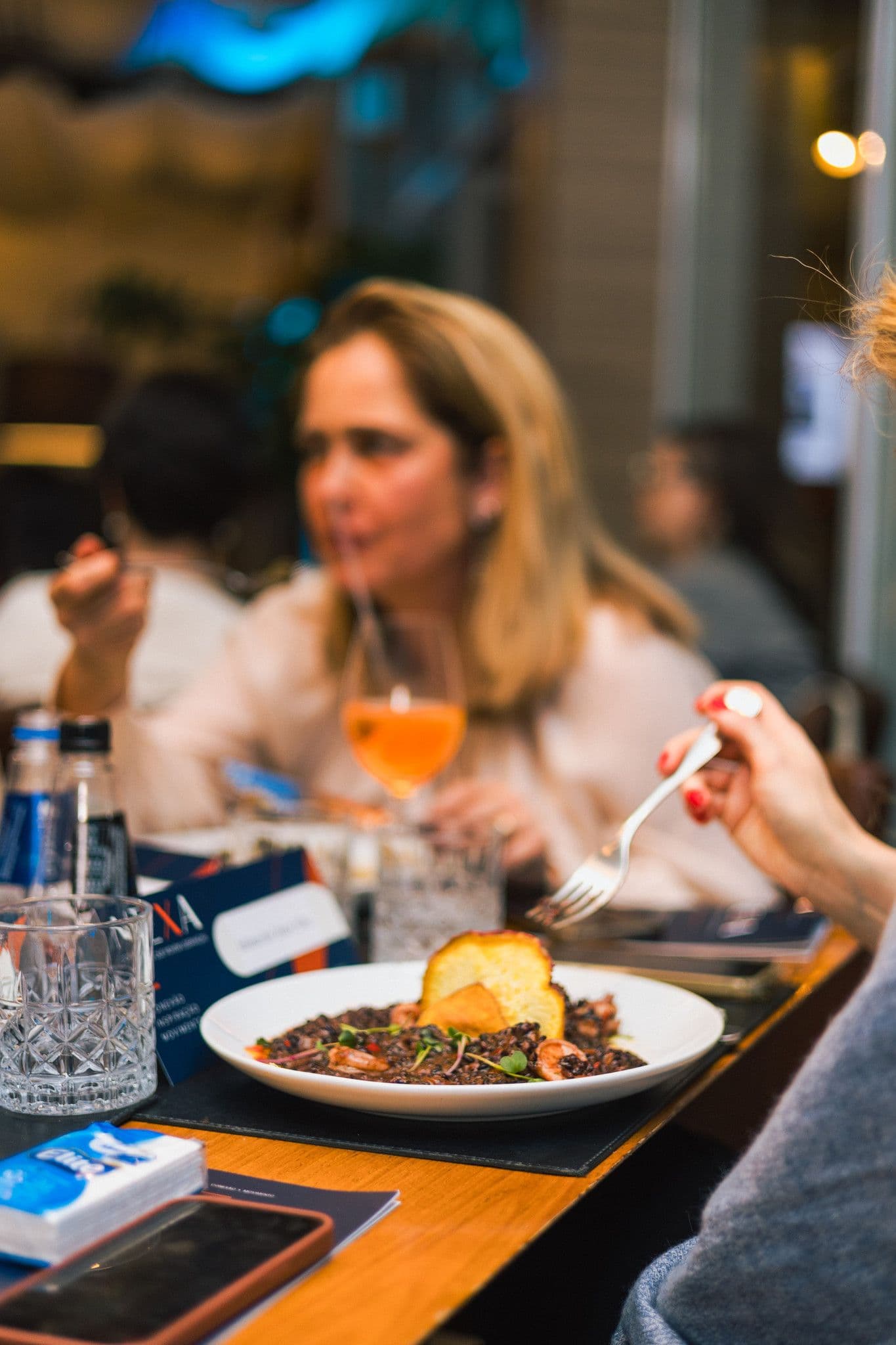 Uma participante sorri à mesa durante o jantar, com um drinque laranja ao lado do prato.