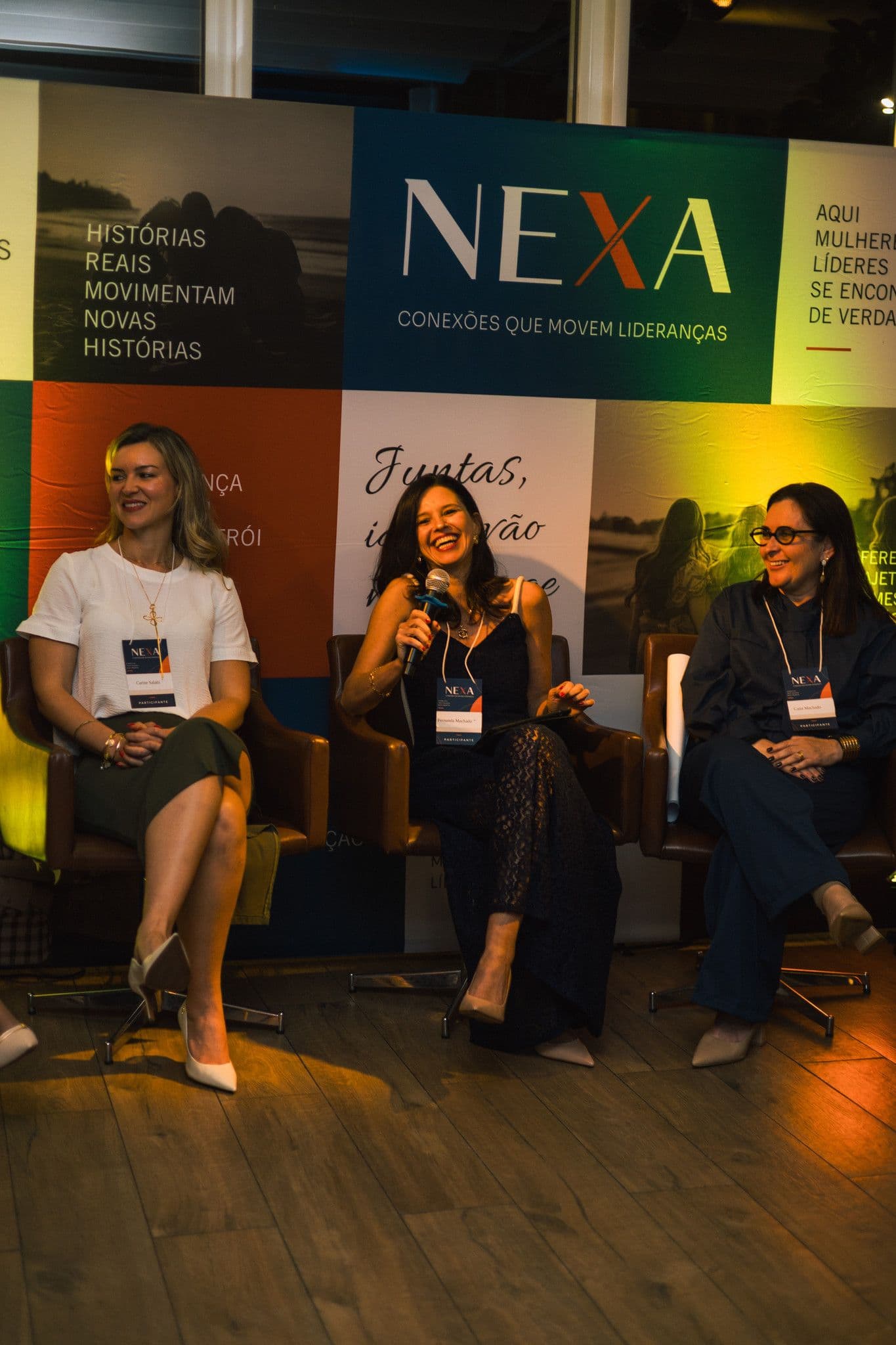 Três mulheres do painel sentadas em poltronas; a do meio fala ao microfone e as outras escutam.