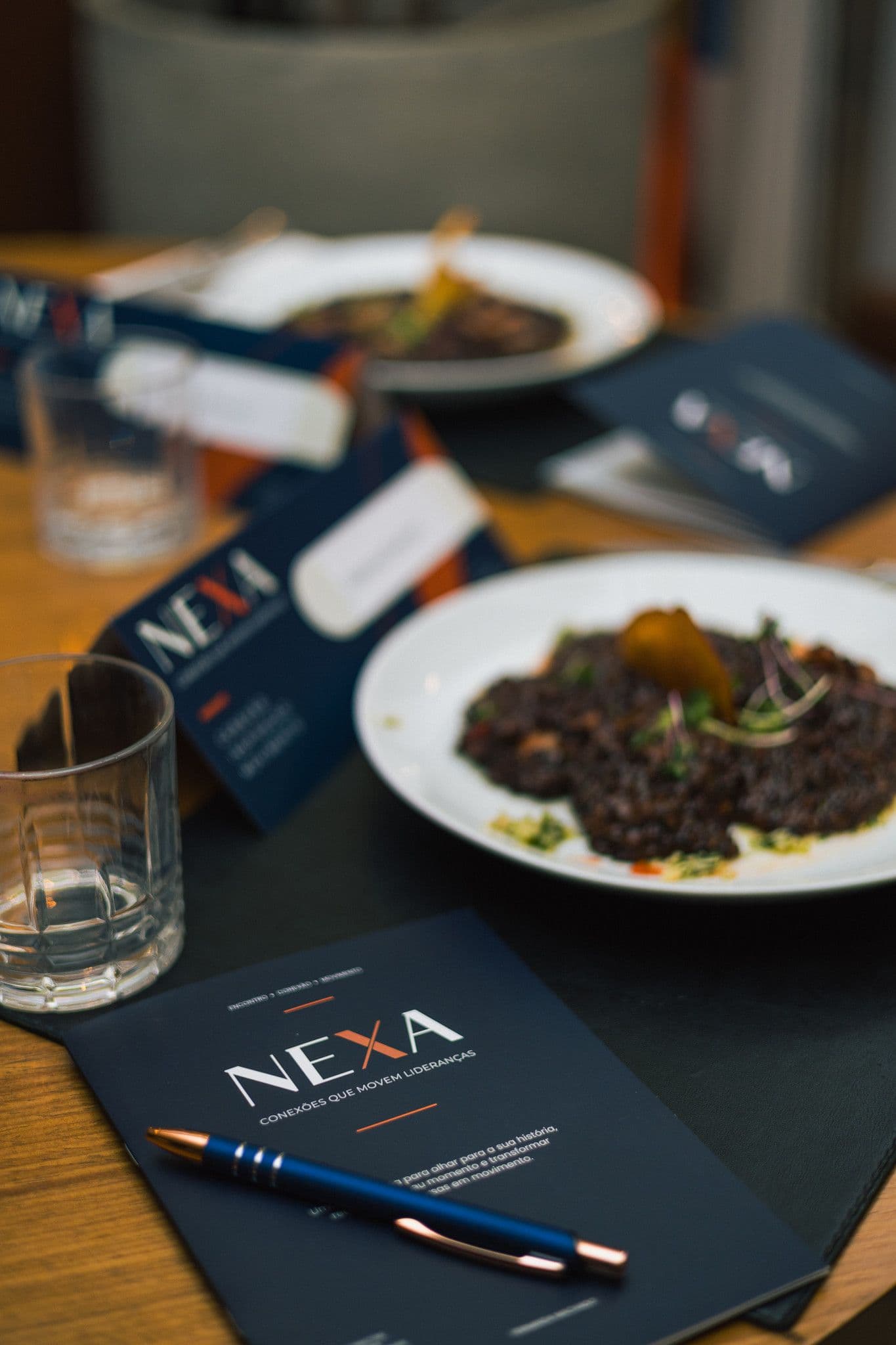 Prato de arroz negro sobre a mesa, com o caderno da NEXA e uma caneta em primeiro plano.