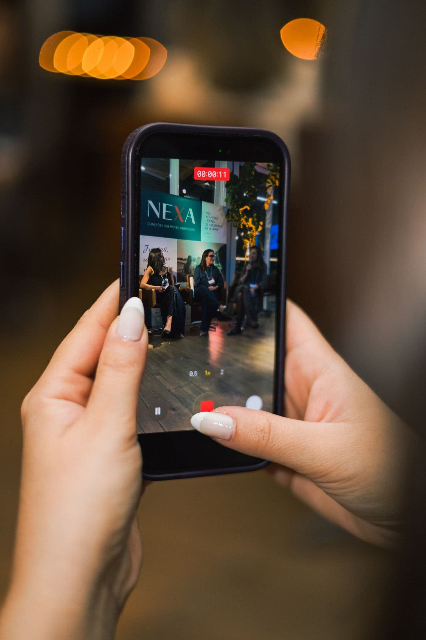 Mãos seguram um celular que filma o painel, com luzes desfocadas ao fundo.