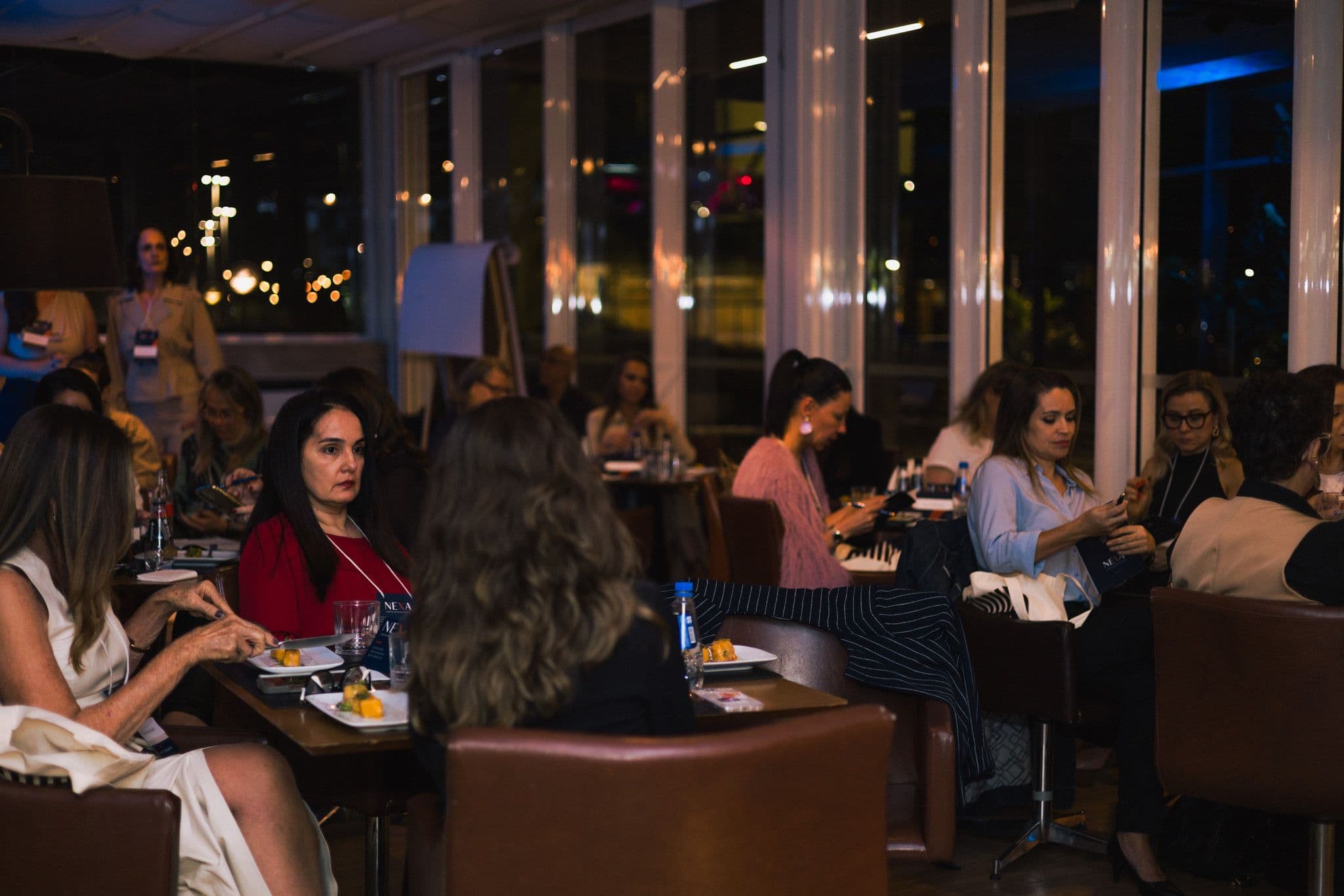 O salão visto do fundo: mesas cheias, conversa entre as participantes, noite lá fora.