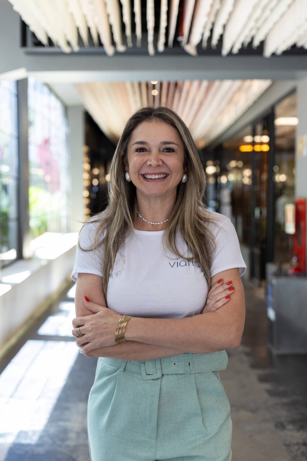 Mulher de pé, braços cruzados, camiseta branca e calça verde-clara, num corredor de teto de ripas claras.