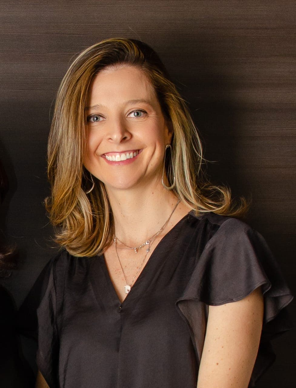 Mulher sorrindo, cabelo castanho-claro na altura dos ombros e blusa preta de decote em V, diante de parede escura.