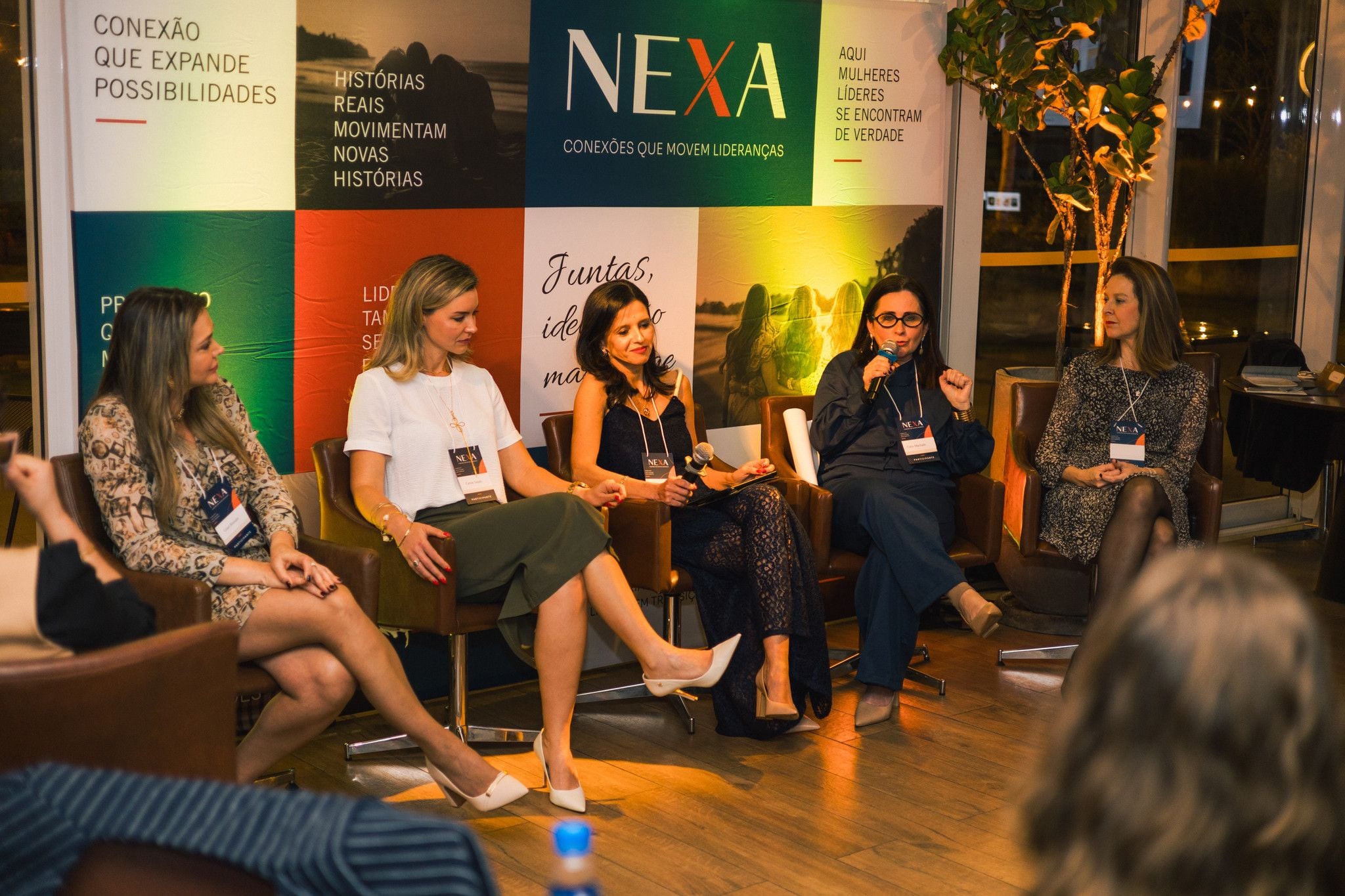 As convidadas do painel sentadas em roda, em poltronas de couro, diante do painel NEXA, com o público à frente.