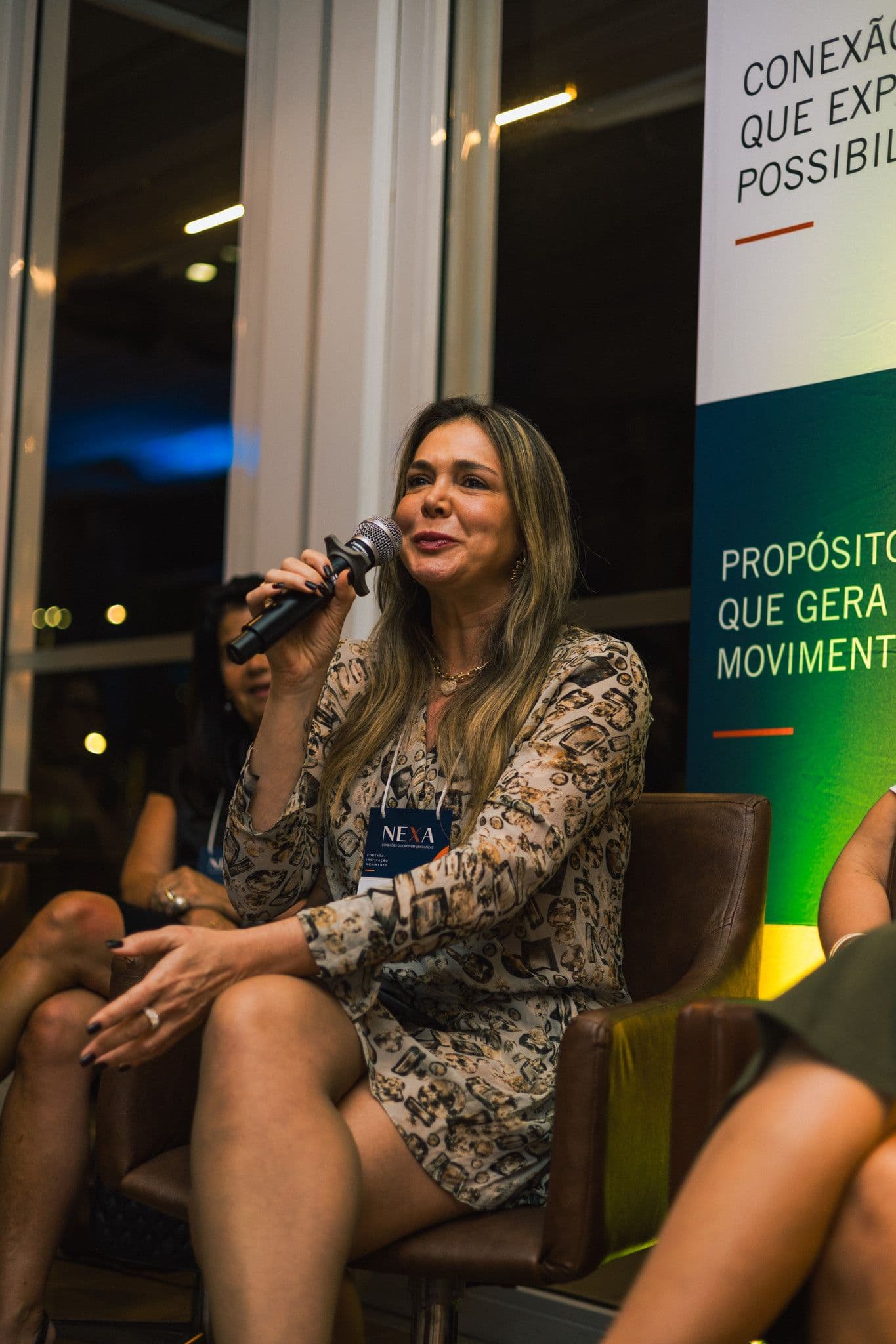 Uma convidada do painel, de vestido estampado, sorri enquanto fala ao microfone, sentada.