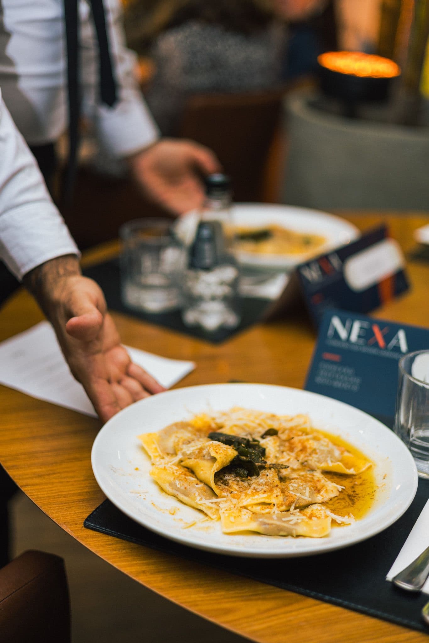 Prato principal servido à mesa: massa recheada com molho dourado, ao lado do cardápio da NEXA.