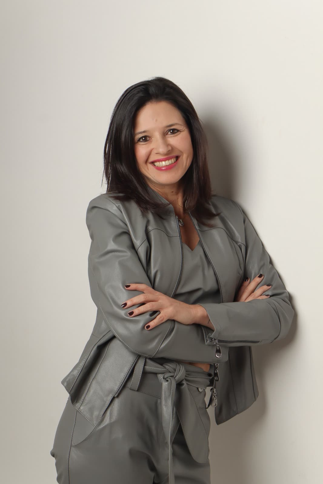 Fernanda Machado em retrato de estúdio, sorrindo, braços cruzados, de jaqueta cinza, encostada numa parede clara.